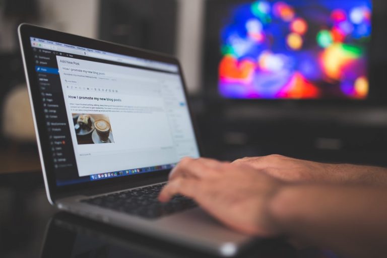 A person is typing on a laptop, which displays a WordPress editor with a draft blog post about promoting blog posts. A blurred, colorful television screen is visible in the background, suggesting they might need resources like SiteGround for hosting and enhancing their WordPress site.