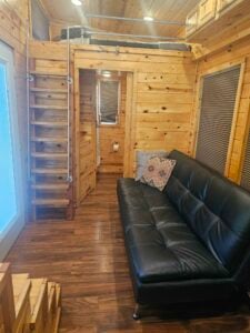 A cozy tiny house interior with wooden walls and flooring. A black leather futon sits on the right, and a ladder leads up to a loft bed. There's a turquoise and pink patterned pillow on the futon. A small doorway leads to the bathroom, and a window is on the left.