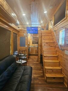 A cozy tiny house interior with wooden walls and ceiling. A black leather sofa sits on the left, facing a wall-mounted TV above the stairs leading to a loft. The compact kitchen with appliances is at the far end. A small dining table with two chairs is near the sofa.