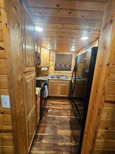 A narrow, wood-paneled kitchen with rustic decor features cabinets, countertops, and walls all made of light-colored wood. It includes a sink, a microwave, an oven, and a refrigerator, all in black stainless steel. Recessed ceiling lights illuminate the space.