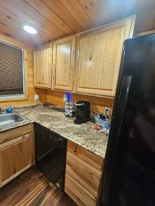 A cozy kitchen with wooden cabinets, a granite countertop, and a black fridge. On the counter, there is a black coffee maker with a K-cup holder, some snacks, a double sink with a window above it, and a black dishwasher below the counter. The floor is dark wood.