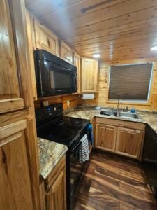 A cozy, rustic kitchen with wooden cabinets and walls. The countertops have a speckled granite finish. The kitchen features a black stove with a microwave above it, a double sink beneath a window with a blind, and a dishwasher. The floor is made of dark wood.