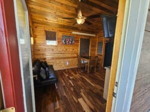 Interior of a rustic cabin with wood-paneled walls and ceiling. Features a small living area with a black sofa, a dining table with four wooden chairs, a ceiling fan, and a wall-mounted TV. Artwork decorates one wall, and natural light enters through a window.