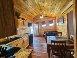 A cozy cabin kitchen and dining area with wooden walls and ceiling. The kitchen has granite countertops and wooden cabinets. A dining table with four chairs is nearby, and a windowed door leads outside. A ceiling fan and a sofa are visible in the background.