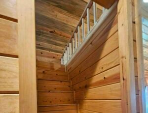 A cozy wooden cabin interior featuring natural wood panels on the walls and ceiling. A wooden staircase with railings leads to an upper level, showcasing the rustic charm and craftsmanship of the space.