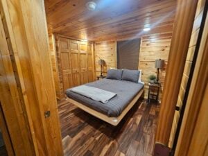 A cozy wooden cabin bedroom features a bed with gray bedding and pillows, illuminated by two side lamps on nightstands. The walls and closet doors are made of wood, creating a warm, rustic atmosphere. A folded blanket lies at the foot of the bed.
