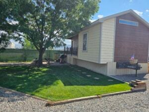 A small yellow house with a wooden accent wall has a well-maintained lawn and a tree casting shade. There's a picnic table and a barbecue grill in the yard. The house number "76" is visible, along with an American flag decoration and some stone steps leading to the porch.
