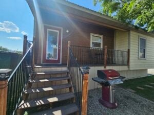 A small house with a wooden porch and steps leading up to a glass front door. The porch is bordered by a black metal railing. A red barbecue grill is positioned on a gravel area beside the steps. The sky is clear and sunny, with a few scattered clouds.