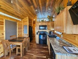 A cozy cabin kitchen with wooden walls and ceiling. The kitchen has black appliances, including a refrigerator, microwave, and oven, as well as a granite countertop. A small wooden dining table with four chairs is near the refrigerator. Plants decorate the space.