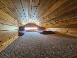 A cozy attic room with wooden walls and ceiling, featuring two mattresses laid out on a carpeted floor, positioned against opposite walls. A small triangular window at the far end lets in natural light, illuminating the space.