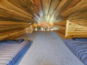 A cozy loft area in a log cabin with wood-paneled walls and ceiling. The space has two twin-sized beds with grey striped blankets on a carpeted floor. A small staircase or ladder leads down to the main area, visible through a large opening in the loft.