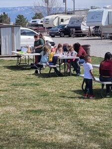 A group of people, including adults and children, gather around tables at an outdoor picnic area with some standing and others sitting. In the background, there are several RVs and a mountain range. The sun is shining, and the grass is green.