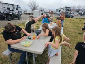 A group of adults and children are sitting at a picnic table outdoors in an RV park, dyeing Easter eggs. Various supplies like containers and bowls with colorful liquid are on the table. Several RVs and a few parked vehicles are visible in the background.