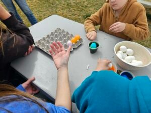Children are gathered around a gray table outdoors, coloring Easter eggs. The table has a carton holding colored eggs, cups with dye, and a bowl of white eggs. One child is holding a pink egg while others are busy with the dyeing process.