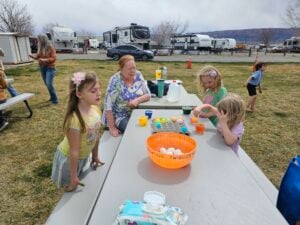 Three children and an older woman are sitting at a picnic table outdoors, dyeing Easter eggs. The table is covered with dyeing supplies, including cups and a basket of eggs. Several RVs and a person playing in the background can be seen. It is a bright day.