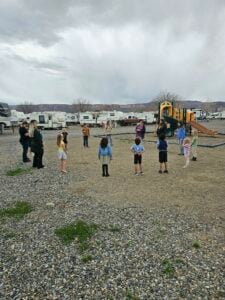 A group of children and adults stand in a circle on a gravel area near a playground. In the background, there are several RVs. The sky is overcast. The children appear to be participating in an outdoor activity led by the adults.