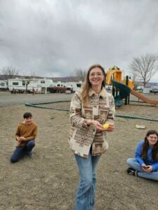 A woman wearing a patterned jacket stands outdoors holding a yellow plastic egg. Behind her, two children are seated on the ground. The area appears to be a playground with RVs and barren trees in the background. The weather is cloudy.