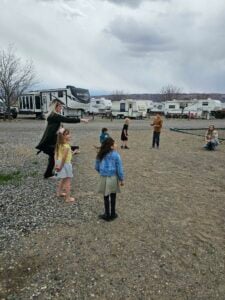 A group of children and adults are playing outside on a gravel and dirt ground near several parked RVs. The sky is overcast. One child is blowing bubbles while others watch and participate. Some adults are standing and observing, while one is crouching down.