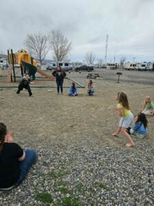 A group of people, including children and adults, are gathered outdoors near a playground. Some children are playing while others sit on the ground. The background includes parked RVs and leafless trees under an overcast sky.