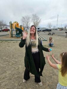 A woman is smiling and waving at the camera while standing in a playground. She has long, light-colored hair and is wearing a dark coat. A child in a light-colored dress is reaching up toward her. In the background are playground equipment, picnic tables, and bare trees.