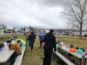 A group of people, including children and adults, are gathered outdoors at picnic tables with RVs and leafless trees in the background. Children are sitting and leaning on the tables, while adults stand and walk nearby, engaging with them.