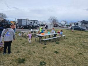 A group of children and adults gather around picnic tables on a grassy area in a campground. Several RVs and trailers are parked in the background. Some children are seated at the tables while others play nearby. The scene appears to be a casual outdoor gathering.