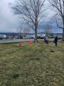 A grassy field with traffic cones set up, a child running towards them, and several people in the background. There are leafless trees and several RVs parked nearby on a gravel surface under a cloudy sky.