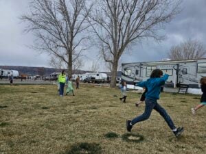 Children are playing and running on a grassy field in front of several RVs and leafless trees. Two adults stand watching them. Dark, cloudy skies are overhead.