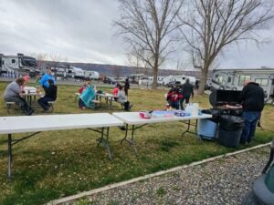 A group of people is gathered outdoors at a campground with RVs in the background. Some are sitting at picnic tables eating, while others are standing near a person grilling food. The sky is overcast, and the area is grassy with bare trees.