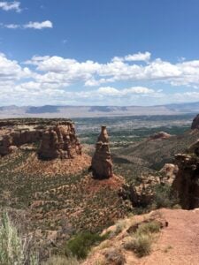 A scenic view of the Colorado National Monument featuring a tall, narrow rock formation in the center with rugged cliffs on the sides. The landscape extends to a distant valley and mountain range under a blue sky with scattered clouds. Vegetation covers the foreground.