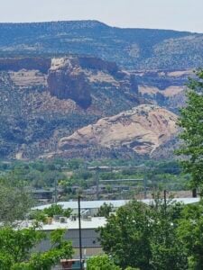 A scenic view featuring a mountainous landscape with rugged cliffs and formations in the background. In the foreground, a small town with buildings and greenery is visible. Trees partially frame the scene, adding a natural element to the composition.