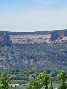 A scenic view of a cliff with layered rock formations in the background, a blue crane visible in the midground, and green vegetation in the foreground under a clear blue sky.