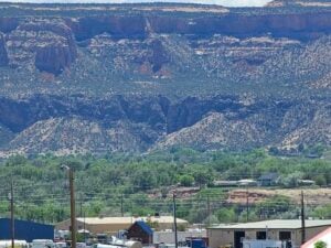 A landscape photograph featuring a rocky, mountainous terrain in the background with layers of ridges. In the foreground, there is a small town with various buildings, vehicles, and greenery, including trees and bushes dotted across the landscape.