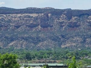 A vast landscape view of rugged cliffs and mesas with layered rock formations in the background, covered sparsely with vegetation. In the foreground, lush green trees and a few buildings can be seen. The sky is clear and blue.