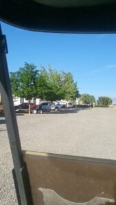 A parking lot with several vehicles, including a pickup truck hauling a trailer, on a clear sunny day. Trees with green foliage line the background along with a white building. The foreground appears to be viewed from within a vehicle.