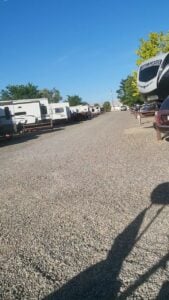 A gravel road lined with parked RVs and trailers on both sides. The sky is clear and blue, and trees with green leaves are visible in the background. Shadows of nearby objects fall across the gravel.