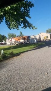 Parked RVs and trailers in a campground with clear skies and green trees in the background. Gravel pathway leads to the RVs which are situated on grassy plots. A tree branch extends over the top left corner of the image, casting light shade.