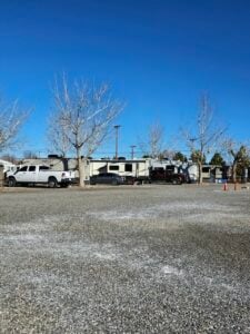 A gravel lot with several recreational vehicles (RVs) and trucks parked under leafless trees. Orange cones are visible on the right, and telephone poles and utility lines can be seen in the background against a clear blue sky.