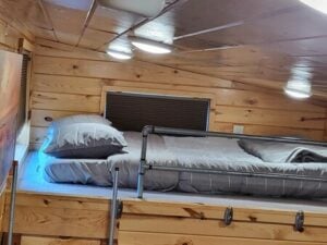 A cozy loft bed with a wooden interior features a neatly made bed with gray sheets, pillows, and a blanket. The space is well-lit with ceiling lights, creating a warm and inviting atmosphere. There is a small window with a closed blind behind the bed.