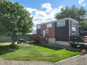 A modern tiny house with brown siding and a small wooden porch is parked on a neatly manicured lawn. There's a large tree providing shade to a wooden picnic table on the left side, and a black pickup truck is parked nearby. A fence and more trees are in the background.