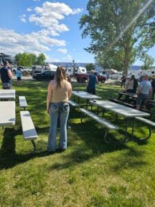 People are gathered outdoors in a grassy park area with picnic tables. Several trailers and RVs are visible in the background. One individual, wearing jeans and a tank top, stands in the foreground, facing away from the camera. It is a sunny day with a clear blue sky.