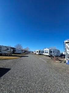 A wide gravel path in an RV park, flanked by numerous parked RVs on both sides. The sky is clear and blue, with a few bare trees visible in the distance. The overall scene is bright and calm.
