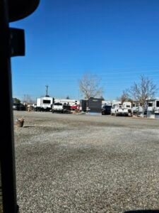 A gravel parking area in a campground with multiple RVs and trailers parked in rows. Several leafless trees and potted plants are scattered around the area under a clear blue sky. A few people and vehicles are visible among the RVs.