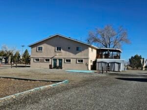 A two-story, beige building with a spacious gravel yard under a clear blue sky. The structure has several windows and a small wooden staircase leading to a balcony on the right side. Sparse trees and additional small structures are visible in the background.