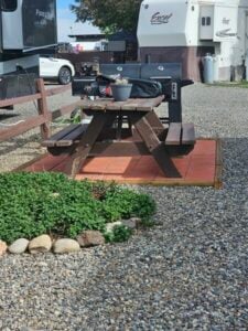 A wooden picnic table on a brick patio surrounded by gravel. The table is next to an RV with a grill on one side and some potted plants on top. There is a small garden bed with green plants and a border of rocks in front of the patio.