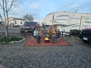A man and a woman sit in foldable chairs around a small fire pit on a paved area at a campsite. Behind them are a large RV and a parked Jeep. The sky is partly cloudy, and the campsite is surrounded by gravel and sparse trees.