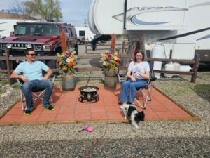 A man and a woman sit in camping chairs on a small tiled patio in front of an RV. They are smiling and looking at a small black and white dog standing near them. The patio is decorated with flowers in metal vases, and a portable fire pit is in the center.