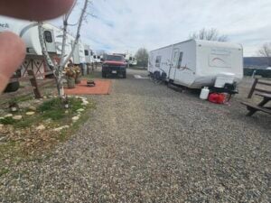 A campground with several RVs parked on gravel lots. One RV has bicycles and propane tanks beside it. A red truck is parked nearby. The area features a small garden and a picnic table. A finger partially obscures the upper left corner of the image.