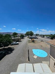 A wide shot of an RV park under a clear blue sky. The image shows several RVs parked along gravel paths with trees providing shade. There is a small fenced playground and a bright blue splash pad in the foreground near a building.