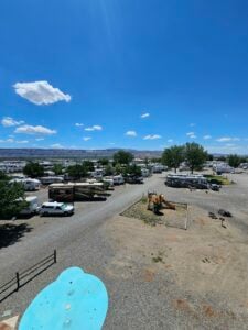 A bird's-eye view of an RV park on a clear, sunny day. The park is filled with various RVs and camping trailers. There is a small playground area with a slide and climbing structure. In the foreground, a dry patch of ground with a disused small pool is visible.